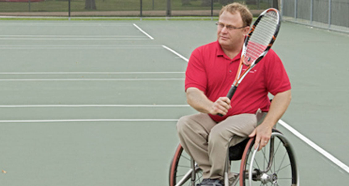 Dr. Cottingham holding a tennis racket sitting on a wheelchair on a tennis court.