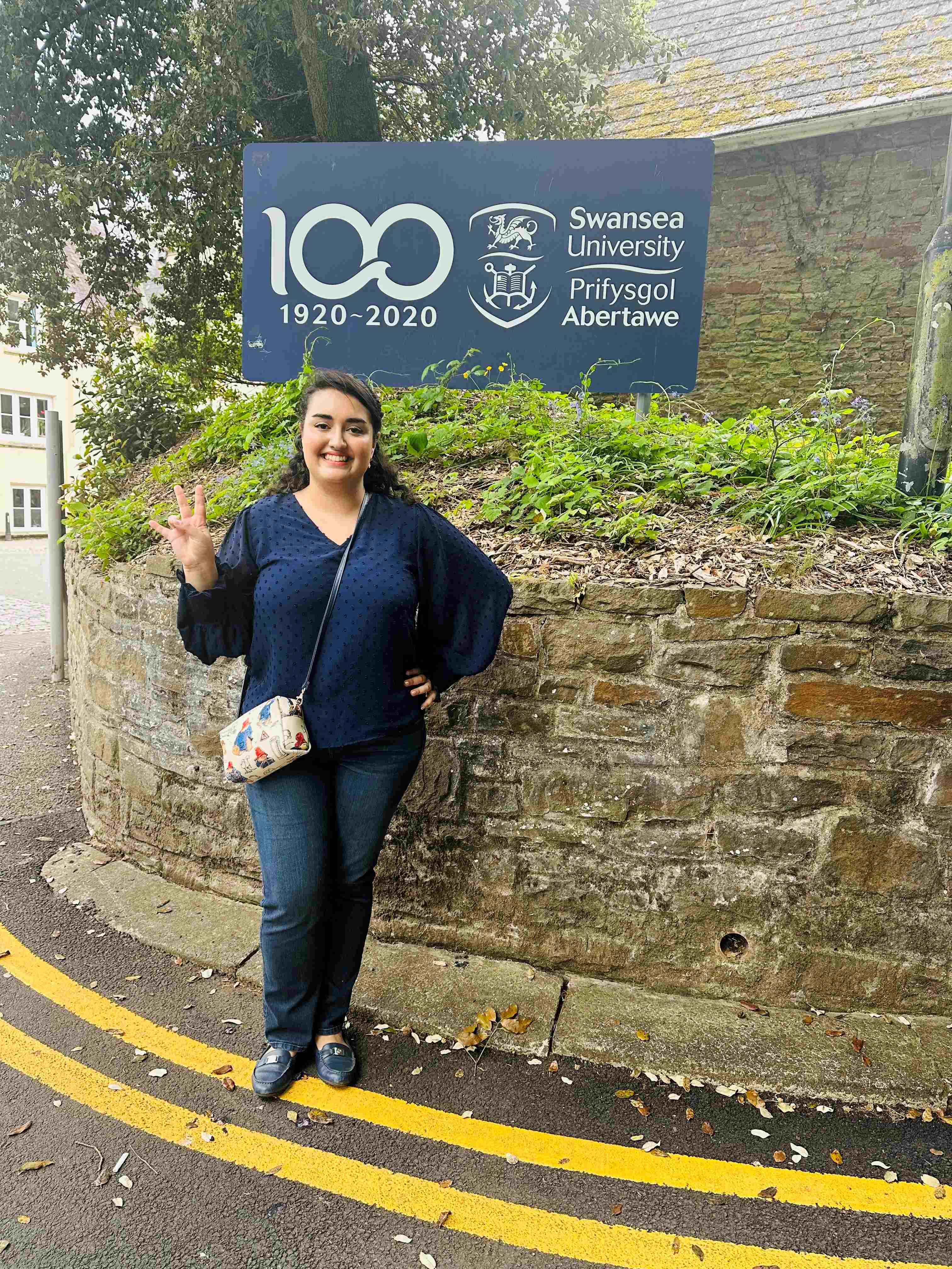Ava standing in front of the Swansea University logo holding up the UH Coogs hand sign.