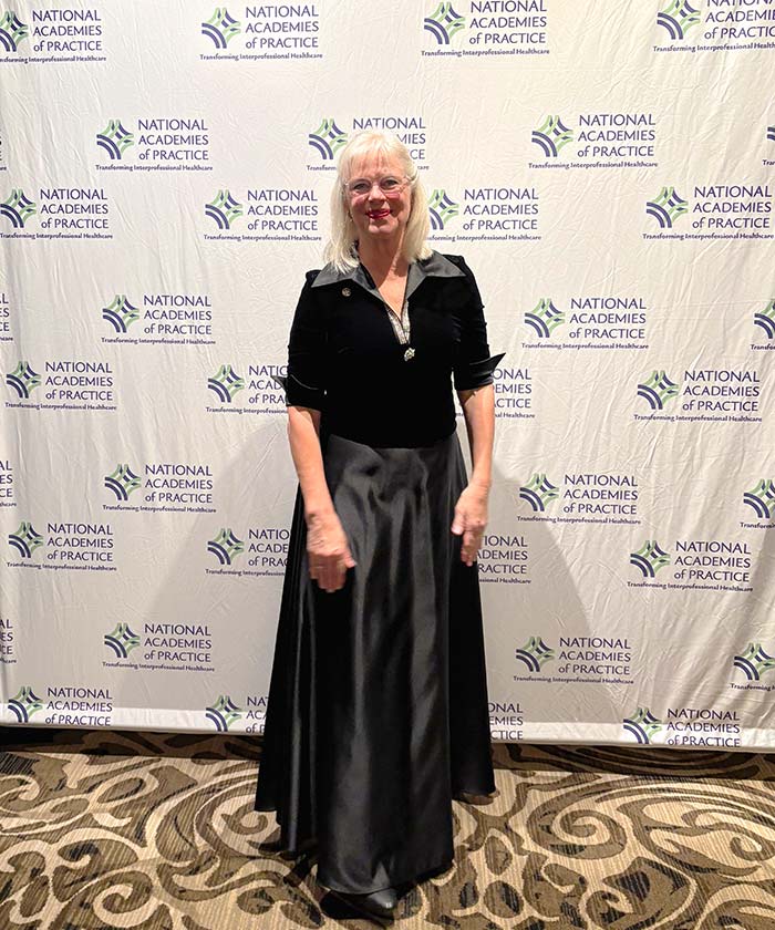 A blonde woman in a black evening gown and jacket smiles in front of a white backdrop with the National Academies of Practice logo.