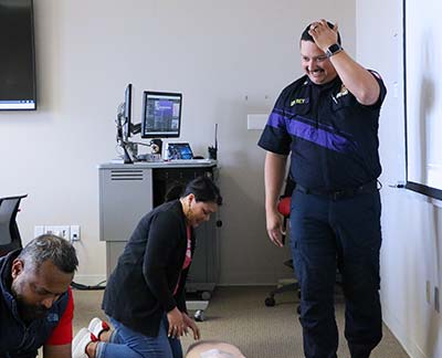 Two people on their knees hunch over CPR training torsos while a Hispanic man in a navy blue Fire Marshal uniform oversees them.