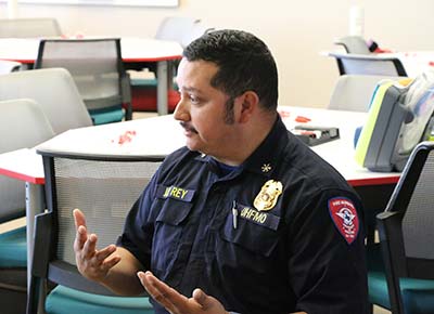 A Hispanic man in a navy blue Fire Marshal uniform faces to the left and gestures with his hands while speaking.